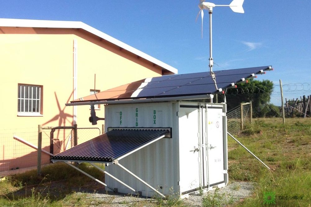 An OffGridBox shipping container in a field with a roof-mounted solar array, wind turbine and solar hot-water tubes, supplying purified water
