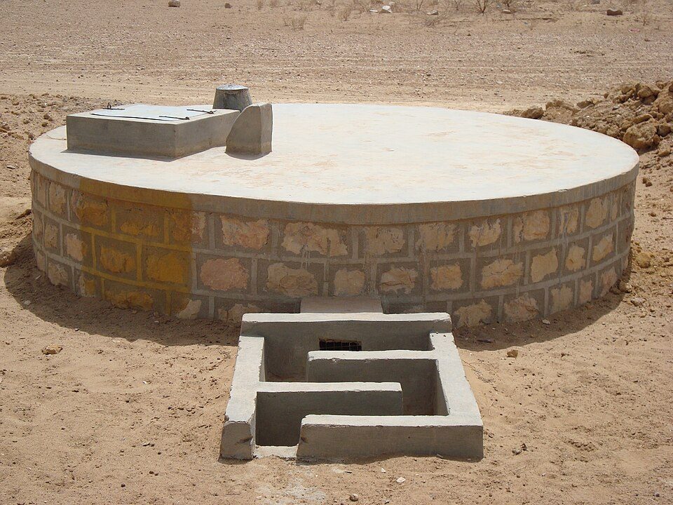 Circular concrete water tank with stone wall and sediment filter box in arid field.