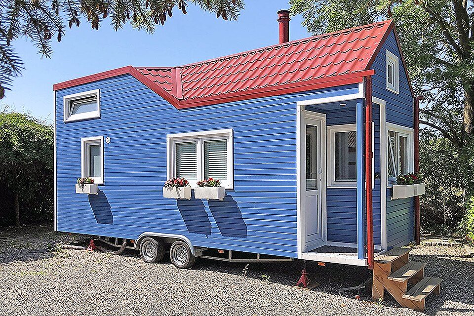 Blue tiny house on trailer with red roof and stove chimney.