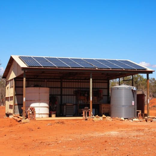 Rural shed with rooftop solar panels and water tanks on red dirt.