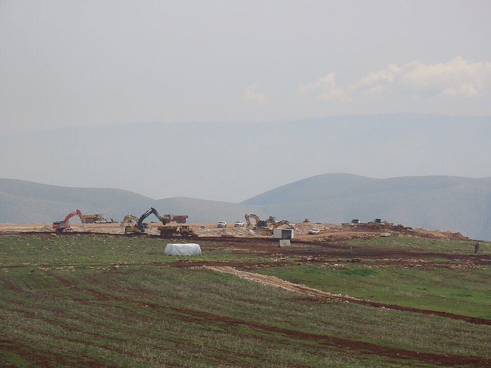 Construction site with excavators on a hillside above a green field.